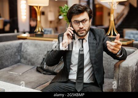 Porträt eines erfolgreichen Geschäftsmannes aus dem Nahen Osten, der mit dem Smartphone spricht, während er in der Hotellobby in einem Sessel sitzt Stockfoto