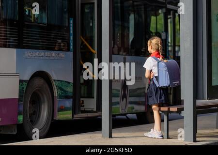 Huzhou, China 22 June 2021: Glückliches kaukasisches Mädchen kehrt in Uniform und roter Krawatte bei Pioneer in die Schule zurück. Am Bahnhof wartet ein Kind auf einen öffentlichen Bus Stockfoto