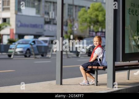 Huzhou, China 22 June 2021: Glückliches kaukasisches Mädchen kehrt in Uniform und roter Krawatte bei Pioneer in die Schule zurück. Am Bahnhof wartet ein Kind auf einen öffentlichen Bus Stockfoto