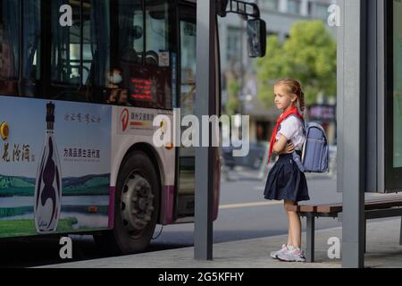 Huzhou, China 22 June 2021: Glückliches kaukasisches Mädchen kehrt in Uniform und roter Krawatte bei Pioneer in die Schule zurück. Am Bahnhof wartet ein Kind auf einen öffentlichen Bus Stockfoto