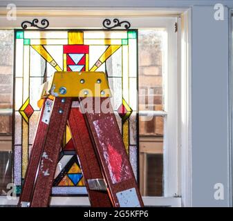 Home Improvement -Hängen Sie ein Art-Deco-Buntglasfenster in einem altmodischen Holzfenster mit einer alten Leiter. Stockfoto