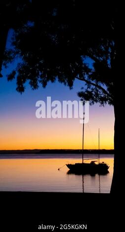Segelboot, das bei Sonnenuntergang vor Anker liegt und durch den Rahmen einer Baumsilhouette vor einem farbenfrohen Himmel betrachtet wird – Platz für Kopien Stockfoto