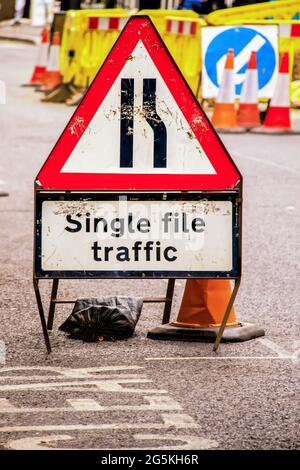 Single File Traffic - altes grungy Beat up Verkehrsschild mit Sandsack auf dem Bürgersteig vor Barrieren und Verkehrskegeln gehalten - Großbritannien - Selektive fo Stockfoto