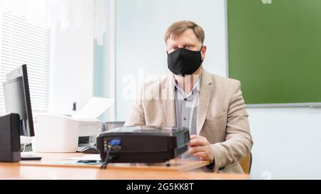 Ein Lehrer sitzt in einem Schulzimmer mit einer schwarzen Schutzmaske an einem Schreibtisch. Stockfoto