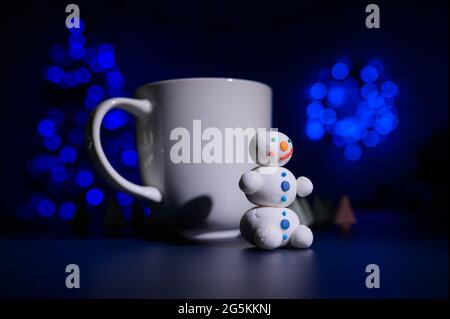 Niedlicher Schneemann und kleine Weihnachtsbäume aus Marschwalben auf dem Hintergrund der Weihnachtslichter. Weißer Becher mit heißer Schokolade und Süßigkeiten. Stockfoto