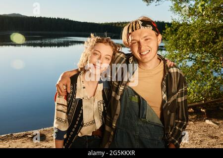 Porträt eines jungen Paares, das die Kamera umarmt und lächelt und im Freien mit einem See im Hintergrund steht Stockfoto