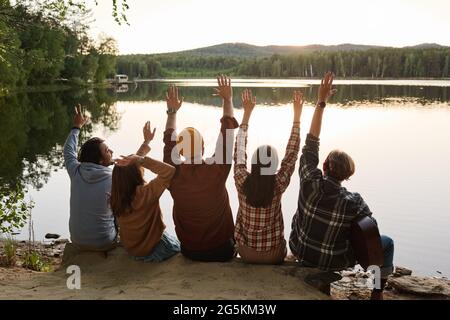 Rückansicht einer Gruppe von Freunden, die ihre Hände hochheben, während sie am Rand des Felsens sitzen und auf den See blicken Stockfoto