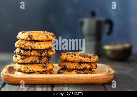 Amerikanische Kekse mit Schokoladenstücken auf einem Holzteller und eine Kaffeekocher auf einem Tisch mit blauem Hintergrund. Hausgemachte Kekse machen, backen Stockfoto