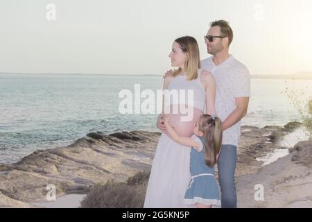 Junge Familie, die mit der Schwangeren am Meer spazieren geht. Die kleine Tochter küsst den Bauch ihrer Mutter Stockfoto