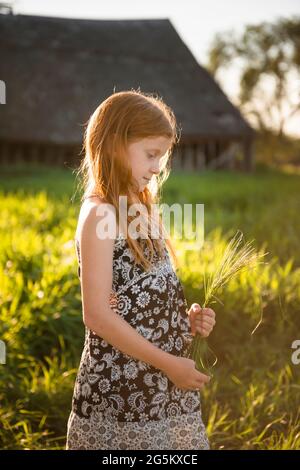 Kleines Mädchen mit roten Haaren im hohen Gras stehend, Scheune im Hintergrund. Stockfoto