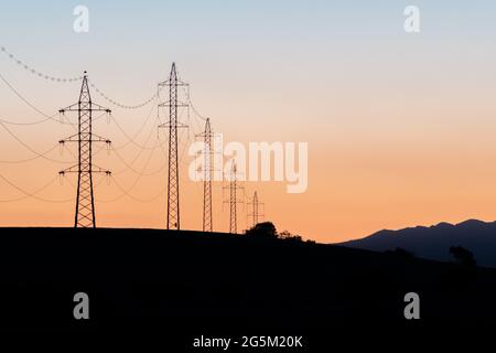Stromtürme über den Bergen bei Sonnenuntergang. Silhouetten von Stromleitungen und Türmen mit einem Storch auf einem der Türme. Stockfoto