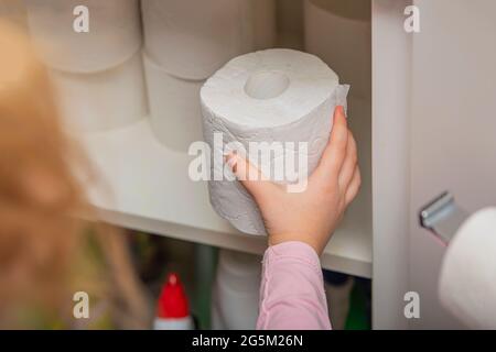 Rolle weißes Toilettenpapier in der Hand. Die Hand des Kindes nimmt eine Rolle Toilettenpapier aus dem Schrank. Viel Toilettenpapier auf dem Hintergrund. Stockfoto