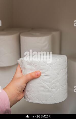Rolle weißes Toilettenpapier in der Hand. Die Hand des Kindes nimmt eine Rolle Toilettenpapier aus dem Schrank. Viel Toilettenpapier auf dem Hintergrund. Stockfoto