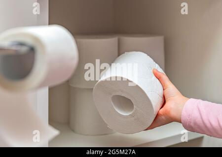 Rolle weißes Toilettenpapier in der Hand. Die Hand des Kindes nimmt eine Rolle Toilettenpapier aus dem Schrank. Viel Toilettenpapier auf dem Hintergrund. Stockfoto