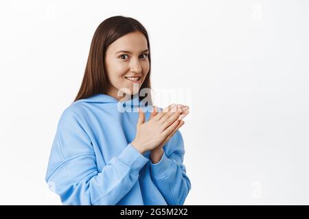 Lächelnde junge Frau versucht, etwas zu versuchen, reiben Hände und beißen Lippe aus Versuchung, Wunsch, leckeres Essen zu probieren, im blauen Hoodie gegen stehen Stockfoto