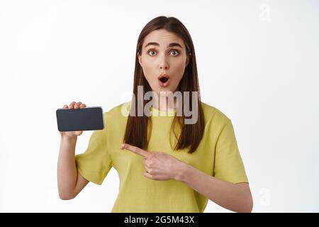 Portrait of surprised girl pointing at horizontal Smartphone screen, keuchen erstaunt, schauen Sie sich tolle neue App, Einkaufen im Online-Shop, stehend Stockfoto