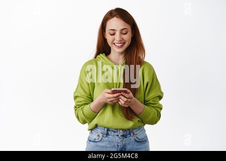 Technologie. Lächelndes rothaariges Mädchen, das auf das Smartphone schaut, am Telefon chattet, die Anwendung auf dem Handy nutzt, im Hoodie vor weißem Hintergrund steht Stockfoto