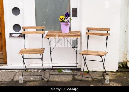 Leerer Holztisch vor dem Restaurant Cafe-Shop mit zwei Stühlen auf der Straße auf dem Bürgersteig und lila gelben Stiefmütterchen Blumen im Blumentopf niemand Stockfoto