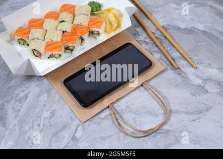 Leckere Sushi-Brötchen und Smartphone auf Steintisch, Draufsicht. Lieferung von Lebensmitteln. Sushi-to-go-Konzept. Stockfoto