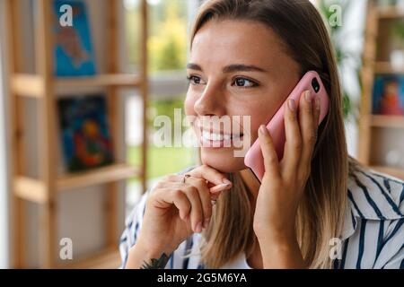 Lächelnd attraktive junge kreative Designerin, die ihr Mobiltelefon nutzt, während sie im Studio arbeitet, aus nächster Nähe Stockfoto