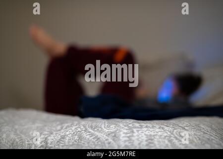 Bokeh verschwommener Hintergrund von dunklem Zimmer und jungen Mädchen auf dem Bett liegend mit Handy in den sozialen Medien und blaues Licht Reflexion auf Gesicht mit b Stockfoto