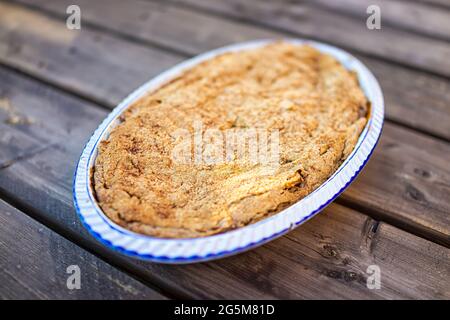 Draußen Holztisch und Nahaufnahme von ganzen Apfelfrucht Kuchen gebackenen Dessert in blauen Keramik-Backform mit Fruchtfüllung und goldene Kruste Stockfoto