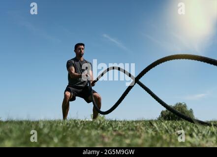 Männlicher Athlet im Freien im Park, Übungen für Ausdauer, mit einem dicken Seil, für Cross-Fitness. Europäisch im Sommer Stockfoto