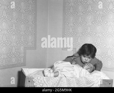 1960s, historisch, in der Ecke eines Zimmers, ein kleines Mädchen, das ihre kleine Schwester überprüft, die auf einem Wurf auf einem Reisebett der damaligen Zeit liegt, England, Großbritannien. Stockfoto