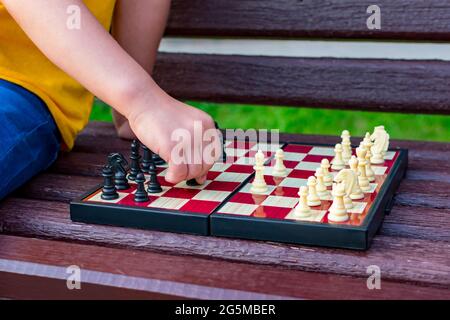 Ein Mädchen spielt Schach im Park auf einer Bank. Schachtag. Die Hand des Kindes ordnet das Schachstück neu. Stockfoto