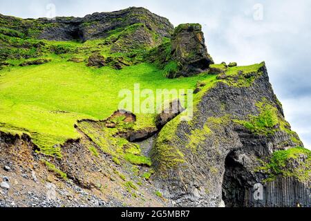 Isländische grüne Wiese Feld Grashügel Berg felsige Klippe Landschaft in Island Sommer in Reynisfjara, Vik mit Basalthöhle Stockfoto