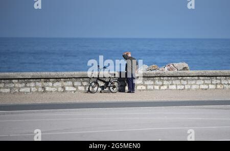Alter grauhaariger Mann mit einem Fahrrad, das auf den Gehwegen steht und an einem sonnigen Tag aufs Meer blickt Stockfoto