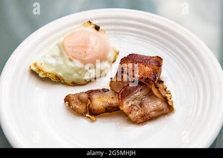 Makro-Nahaufnahme von gekochten, fettigen Speckstreifen und Spiegelei auf weißem Teller beim Frühstücksbrunch, traditionelle amerikanische Mahlzeit Stockfoto