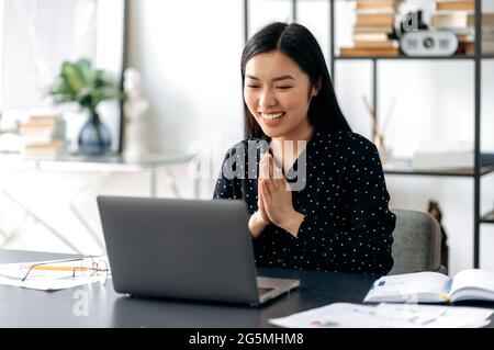 Asiatische glückliche junge Frau, sitzen am Arbeitsplatz, jubeln über Erfolg, Sieg, gutes Ergebnis, Die Handflächen vor ihr zusammengefaltet. Chinesische oder japanische Frau begrüßt den Gesprächspartner während eines Online-Videoanrufs Stockfoto