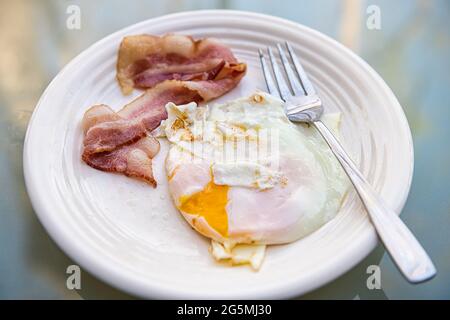Makro-Nahaufnahme von gekochten Speckscheiben und Spiegelei auf weißem Teller beim Frühstücksbrunch, traditionelles amerikanisches Essen und Gabel Stockfoto