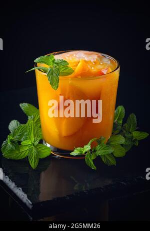 Kalte Getränke im Sommer: Hausgemachte Pfirsich-Sangria mit Eis und Minze im Glas auf schwarzem Hintergrund aus Holz Stockfoto