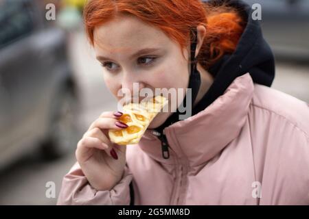 Das Mädchen isst Mehl. Das Mädchen isst auf der Straße einen süßen Kuchen. Leckeres Essen außerhalb des Hauses. Heiße Backwaren in der Hand eines Teenagers. Stockfoto