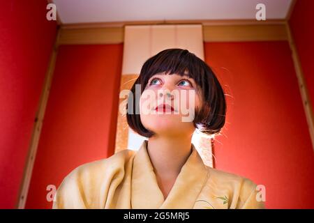 Traditionelles japanisches Haus mit roter Wand auf Nische und hängender Papierrolle mit Frau im Kimono-Kostüm, die mit kurzen schwarzen Haaren aufschaut Stockfoto