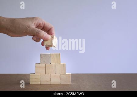 Frau Hand setzen einen quadratischen Holzblock auf Pyramidenstapel von Holzblöcken. Wachstums- oder Erfolgskonzept. Stockfoto