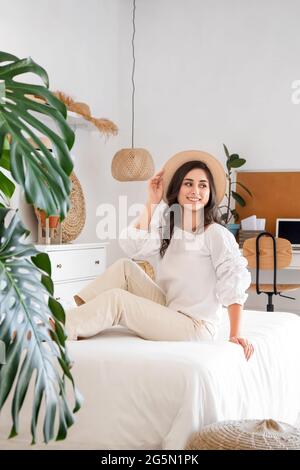 Junge Frau in stilvollem Hut sitzt auf dem Bett im Zimmer Stockfoto