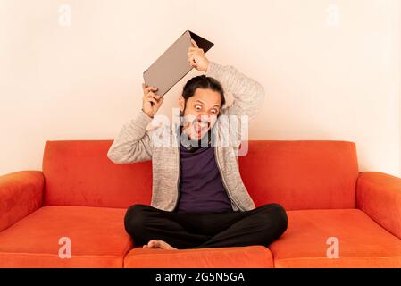Ein junger Mann, der wütend vor Wut den Computer warf und zu Hause im Wohnzimmer auf einem orangefarbenen Sofa saß. Stresskonzept. Stockfoto