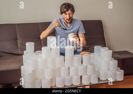 Der Mann isst Joghurt und ist von vielen Joghurtgläsern umgeben, die er in einem Jahr verwendet. Eine riesige Menge von Plastikbehältern für Lebensmittel. Übermäßiger Verbrauch von Stockfoto