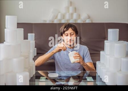 Der Mann isst Joghurt und ist von vielen Joghurtgläsern umgeben, die er in einem Jahr verwendet. Eine riesige Menge von Plastikbehältern für Lebensmittel. Übermäßiger Verbrauch von Stockfoto