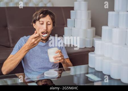 Der Mann isst Joghurt und ist von vielen Joghurtgläsern umgeben, die er in einem Jahr verwendet. Eine riesige Menge von Plastikbehältern für Lebensmittel. Übermäßiger Verbrauch von Stockfoto