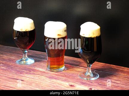 Auf einem Holztisch stehen drei verschiedene Gläser mit einem Drittel verschiedener dunkler Biersorten in einer Reihe. Nahaufnahme. Stockfoto