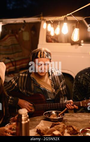 Junger Mann, der für seine Freunde Gitarre spielt, während sie beim Camping im Wald am Tisch sitzen Stockfoto