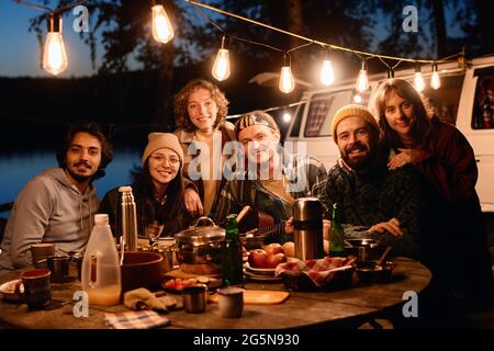 Portrait von glücklichen Freunden, die während des Campens im Wald am Esstisch sitzen und die Kamera anlächeln Stockfoto