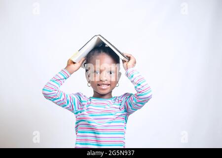 Nettes afrikanisches Mädchen Kind mit gewebter Frisur legt glücklich ein Buch auf ihren Kopf, während sie studiert, um zu zeigen, wie interessant Bildung geworden ist Stockfoto