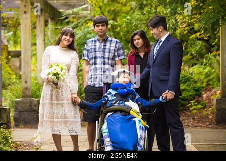 Frisch verheiratete Braut und Bräutigam posiert zusammen mit der Familie im Freien unter weinbedeckter Pergola neben einem kleinen behinderten Jungen im Rollstuhl Stockfoto