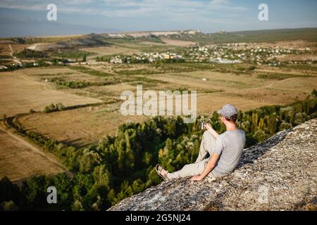 Rückansicht eines jungen Mannes, der Tee oder Kaffee in den Bergen trinkt. Reisender Mann mit Bart hält in den Händen eine Tasse Tee, nach dem Wandern. Reisen, Lifestyle, VA Stockfoto