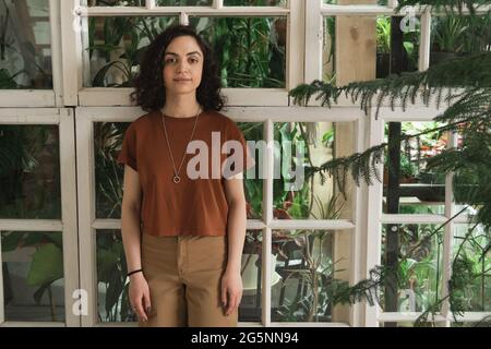 Porträt einer jungen Frau mit dunklen lockigen Haaren, die in einem Blumengeschäft mit Pflanzen und Blumen hinter ihrem Rücken auf die Kamera schaut Stockfoto
